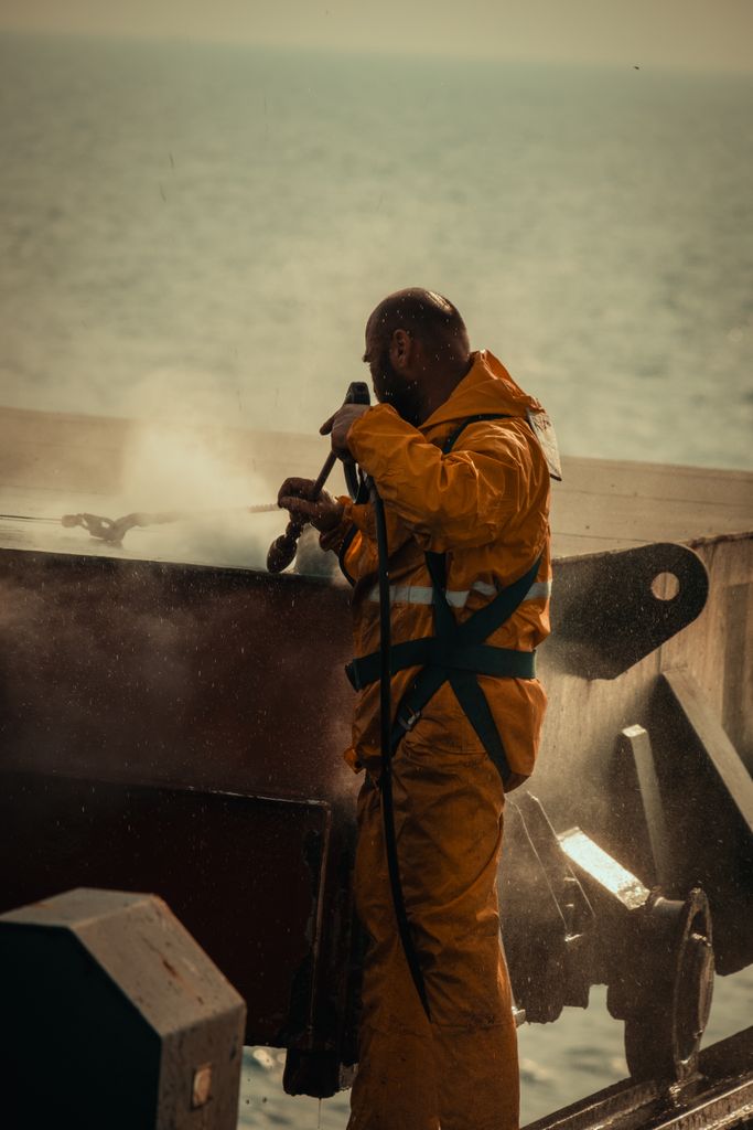 A worker in a yellow rain suit operates equipment on a ship deck with ocean backdrop.